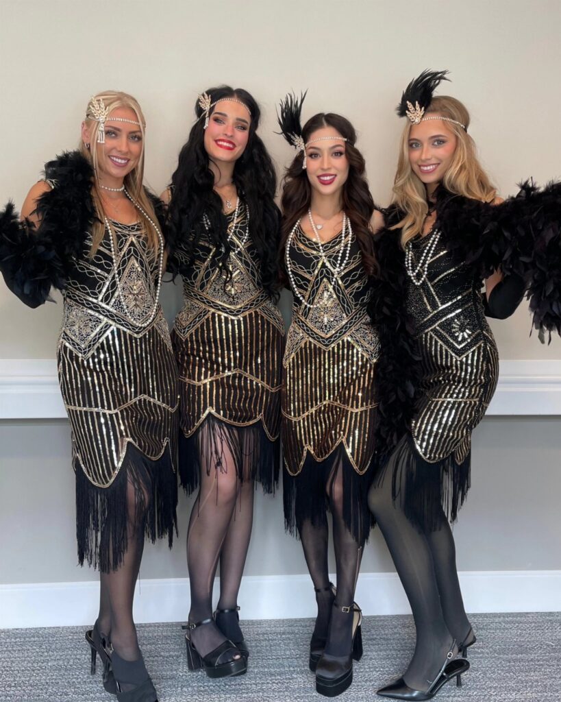 Four women posing in black and gold sequined flapper dresses with fringe and feathers.