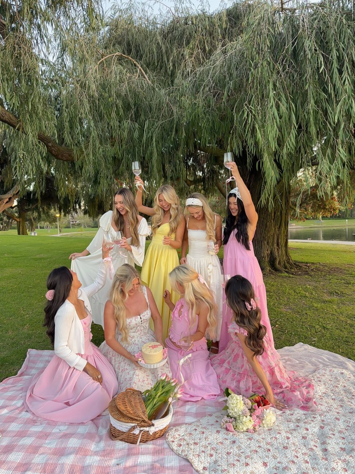 Seven women in pastel dresses sitting on a picnic blanket under a large willow tree.