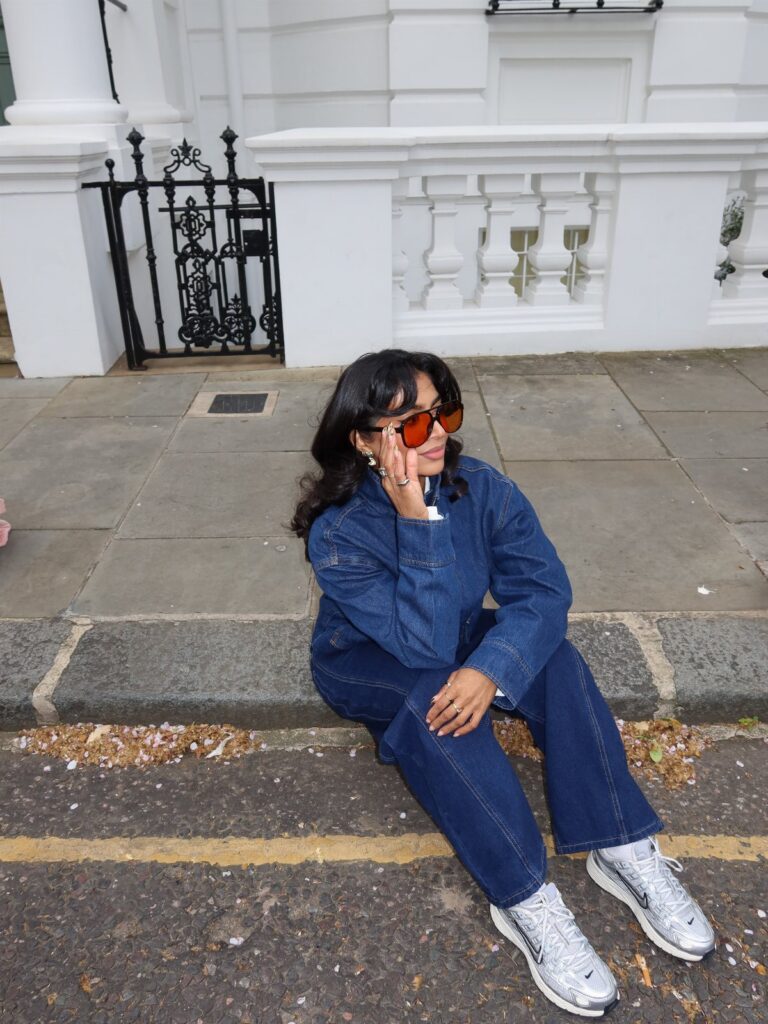 Woman in a denim jacket and denim pants sitting on a sidewalk wearing sunglasses and glittery sneakers.