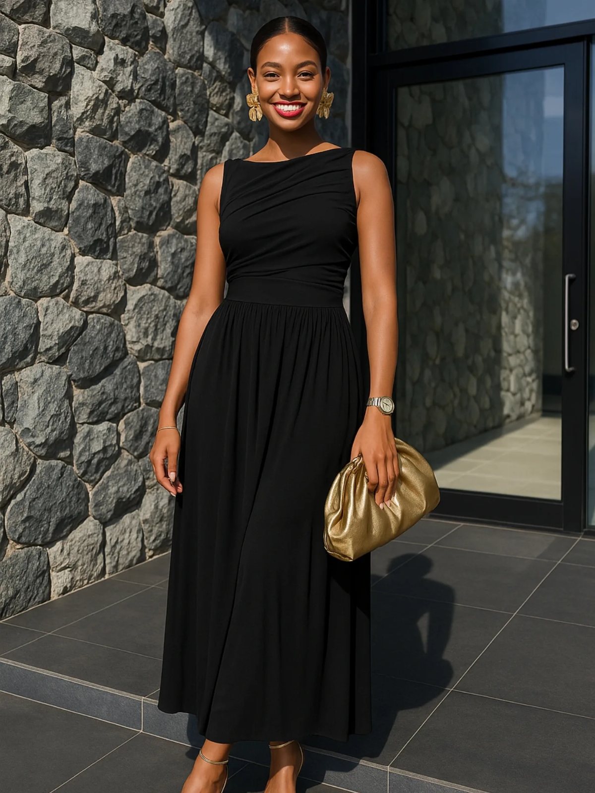 A smiling woman posing in a long, sleeveless black maxi dress and gold accessories.
