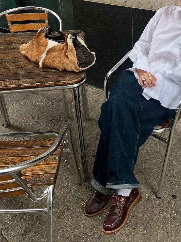Person wearing dark denim jeans and burgundy leather boat shoes sitting at an outdoor cafe table.