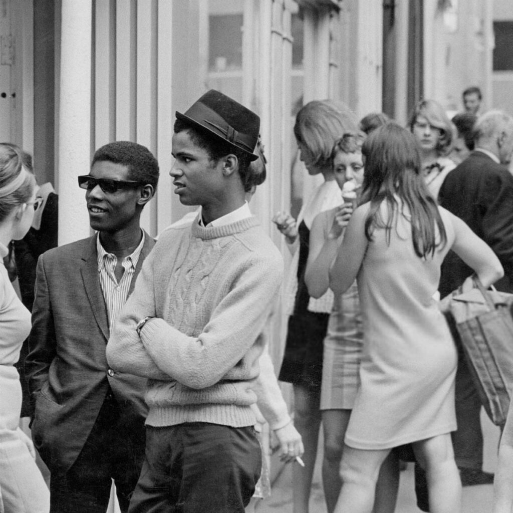 Young people in stylish 1960s attire walking along a busy city street in black and white.