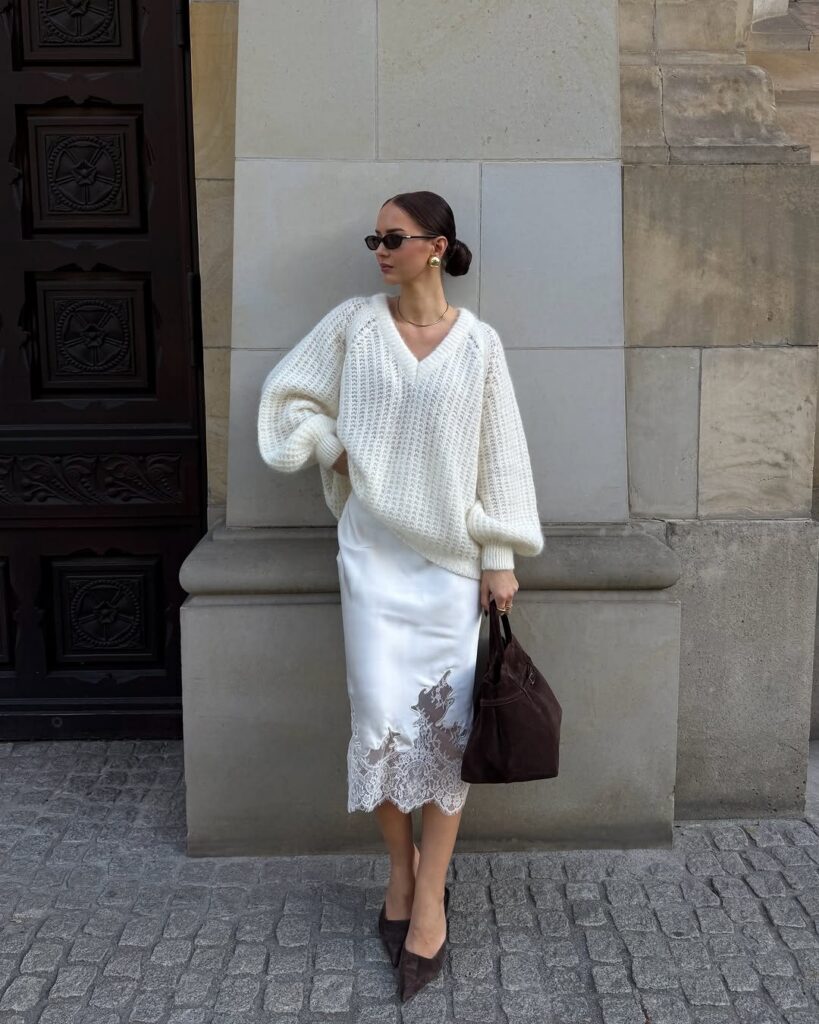 Woman wearing a white cable-knit sweater and a white satin lace skirt standing outdoors.
