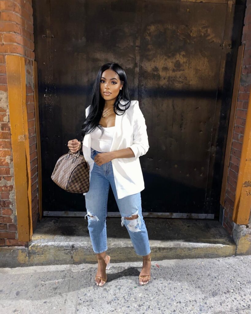Woman in a white blazer, white top, ripped blue jeans, and tan heels holding a handbag.