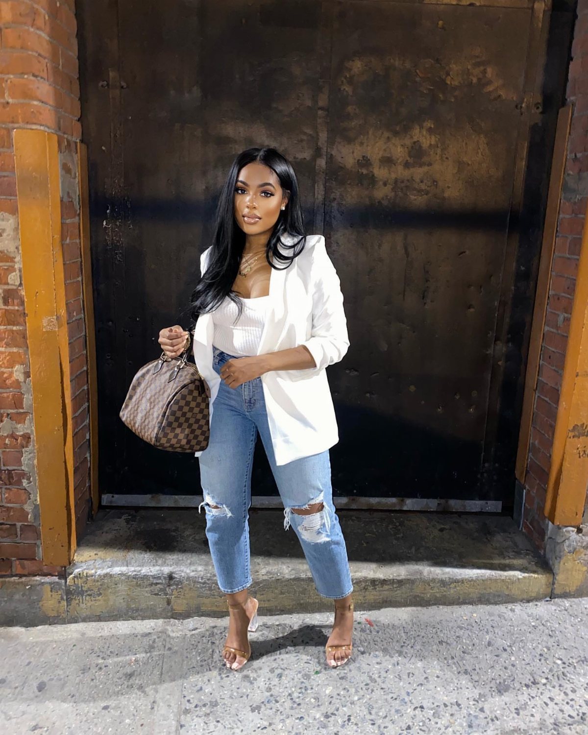 Woman in a white blazer, white top, ripped blue jeans, and tan heels holding a handbag.