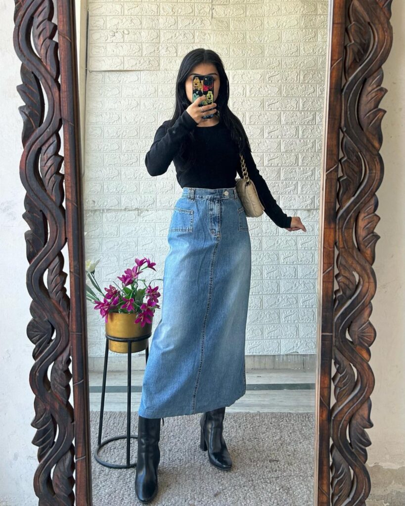 A woman posing in a long denim maxi skirt and black top for a mirror selfie.