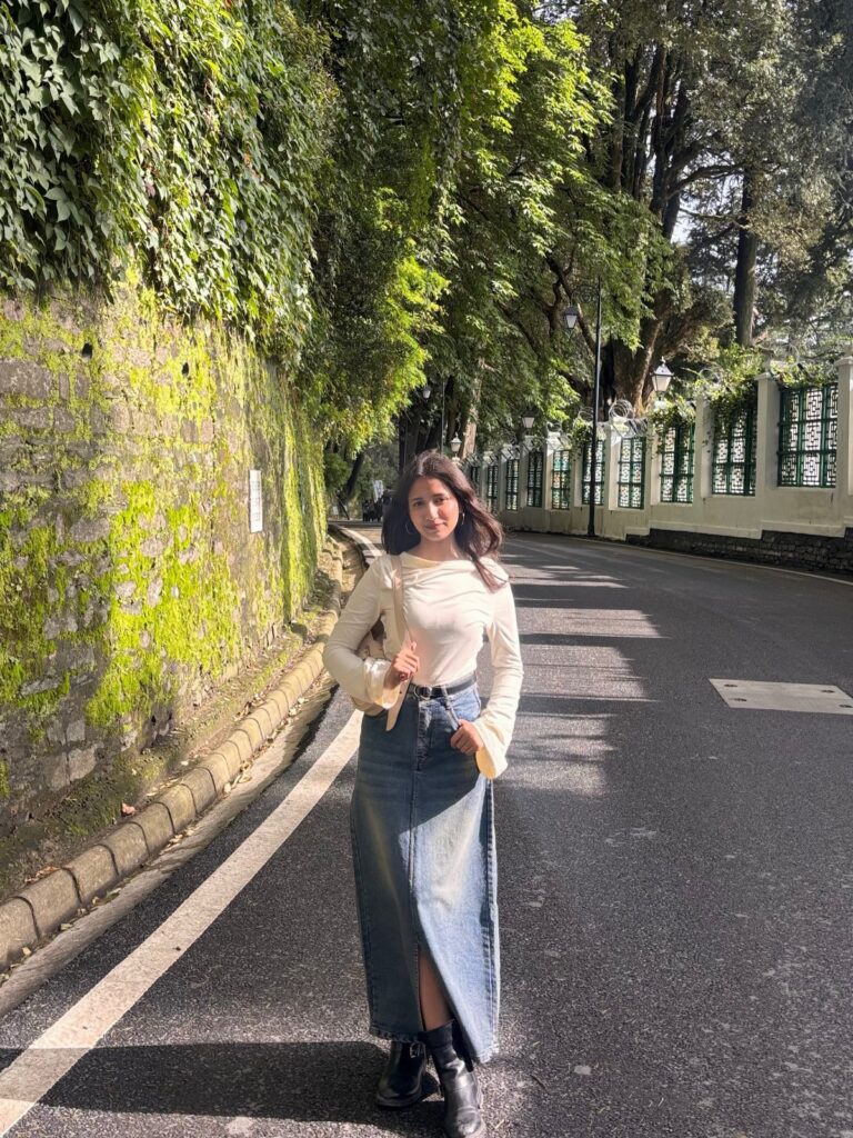 A woman wearing a long denim skirt and cream top standing on a tree-lined road.