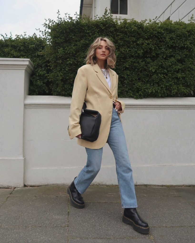 Woman in a beige blazer, blue jeans, and black boots walking past a hedge.