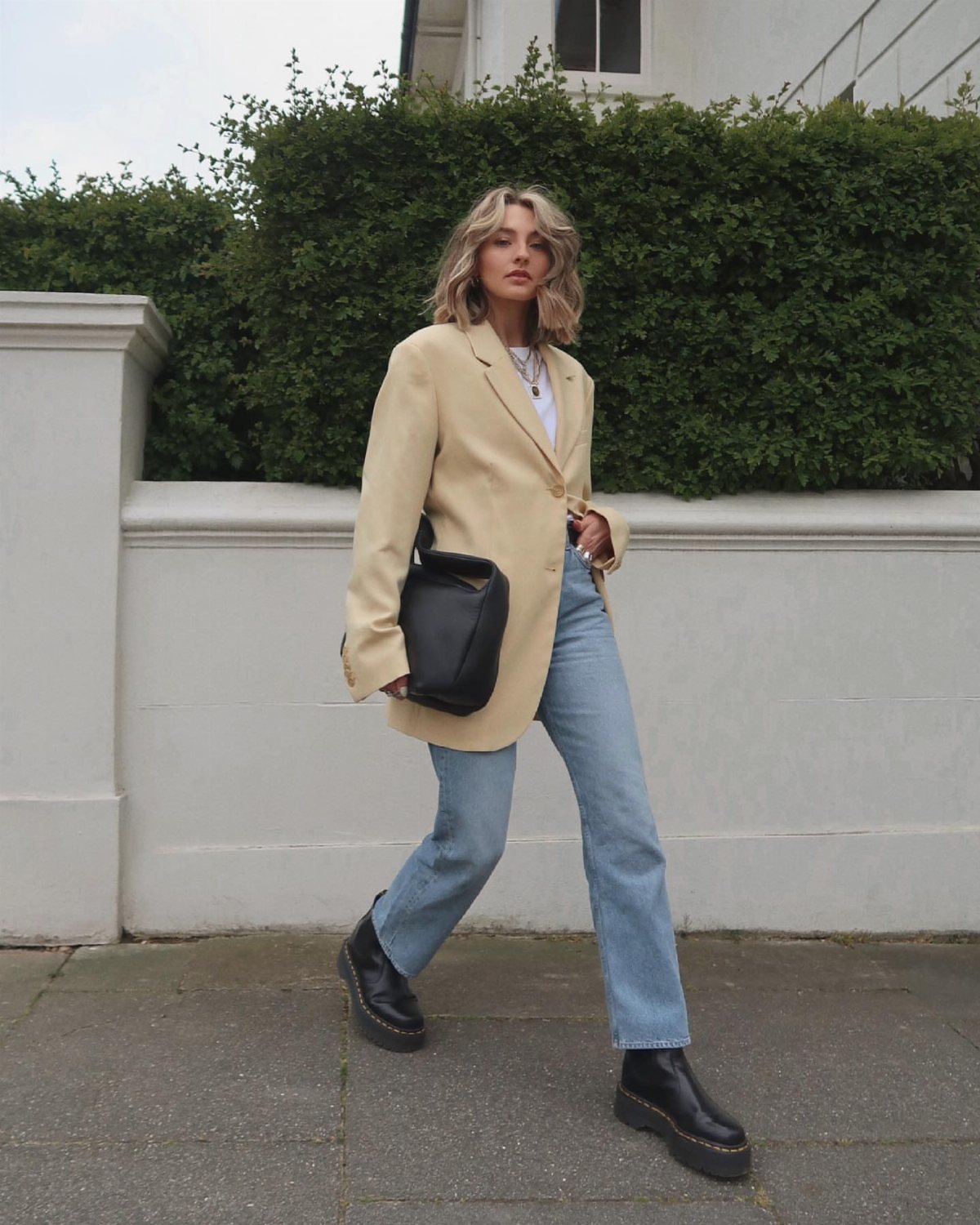 Woman in a beige blazer, blue jeans, and black boots walking past a hedge.