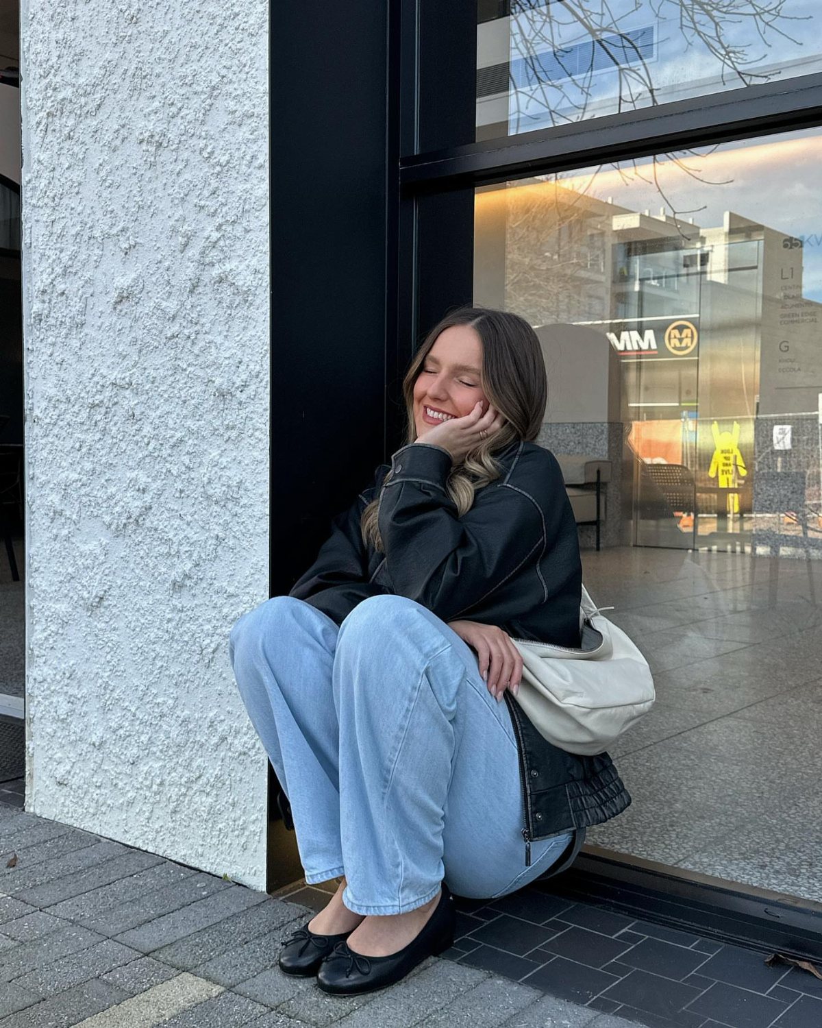 A woman in denim jeans and a black jacket wearing black ballet flats while sitting outdoors.