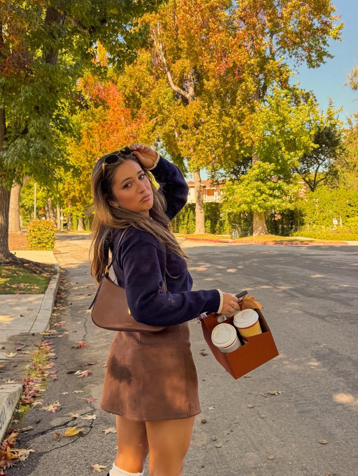 Woman wearing a dark blue sweater and brown skirt standing on a street with autumn trees.