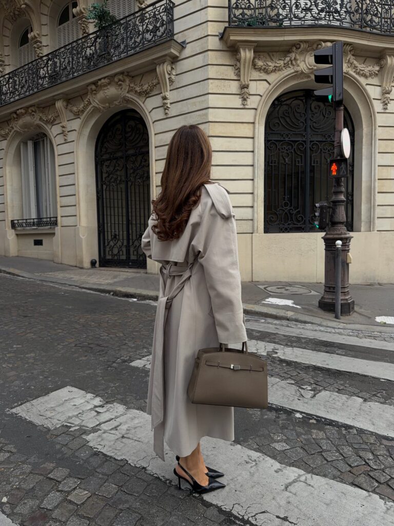 A woman in a long beige trench coat and black slingbacks walking on a city street.