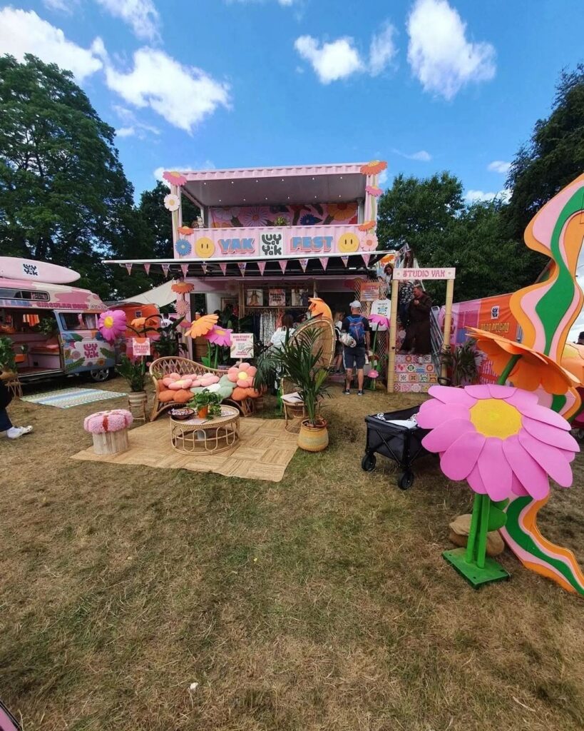A vibrant pink festival pop-up installation with large flowers and lounge seating on a grassy field.