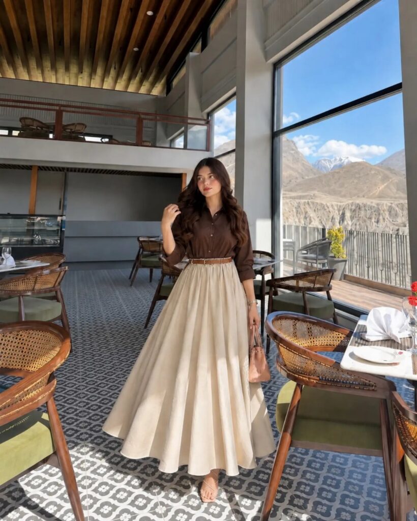 Woman posing in a brown shirt and cream high-waisted maxi skirt inside a cafe.