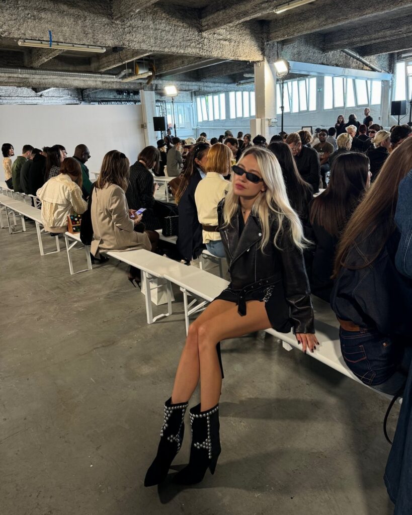 A blonde woman wearing an all-black leather outfit and studded boots sits on a bench.