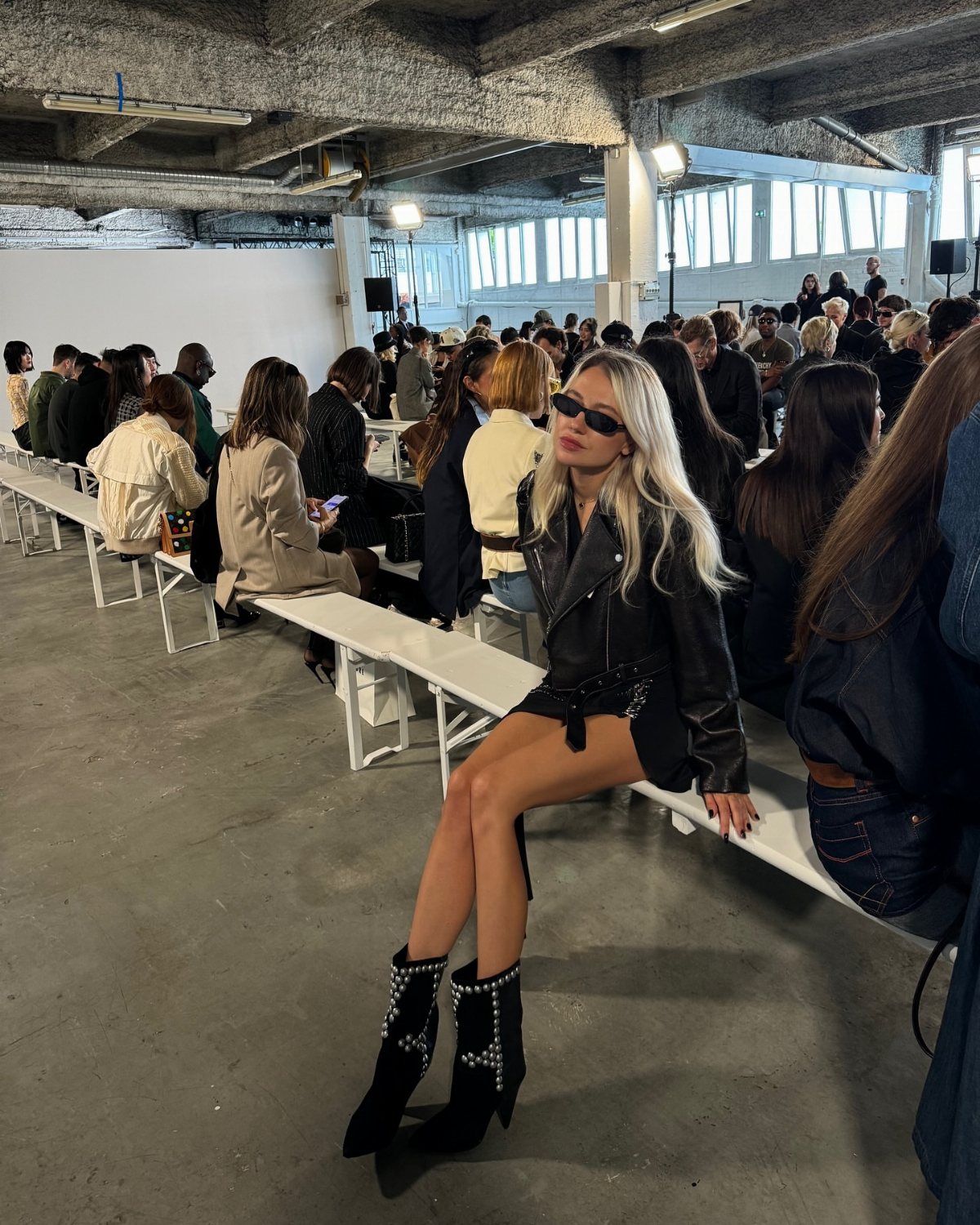 A blonde woman wearing an all-black leather outfit and studded boots sits on a bench.