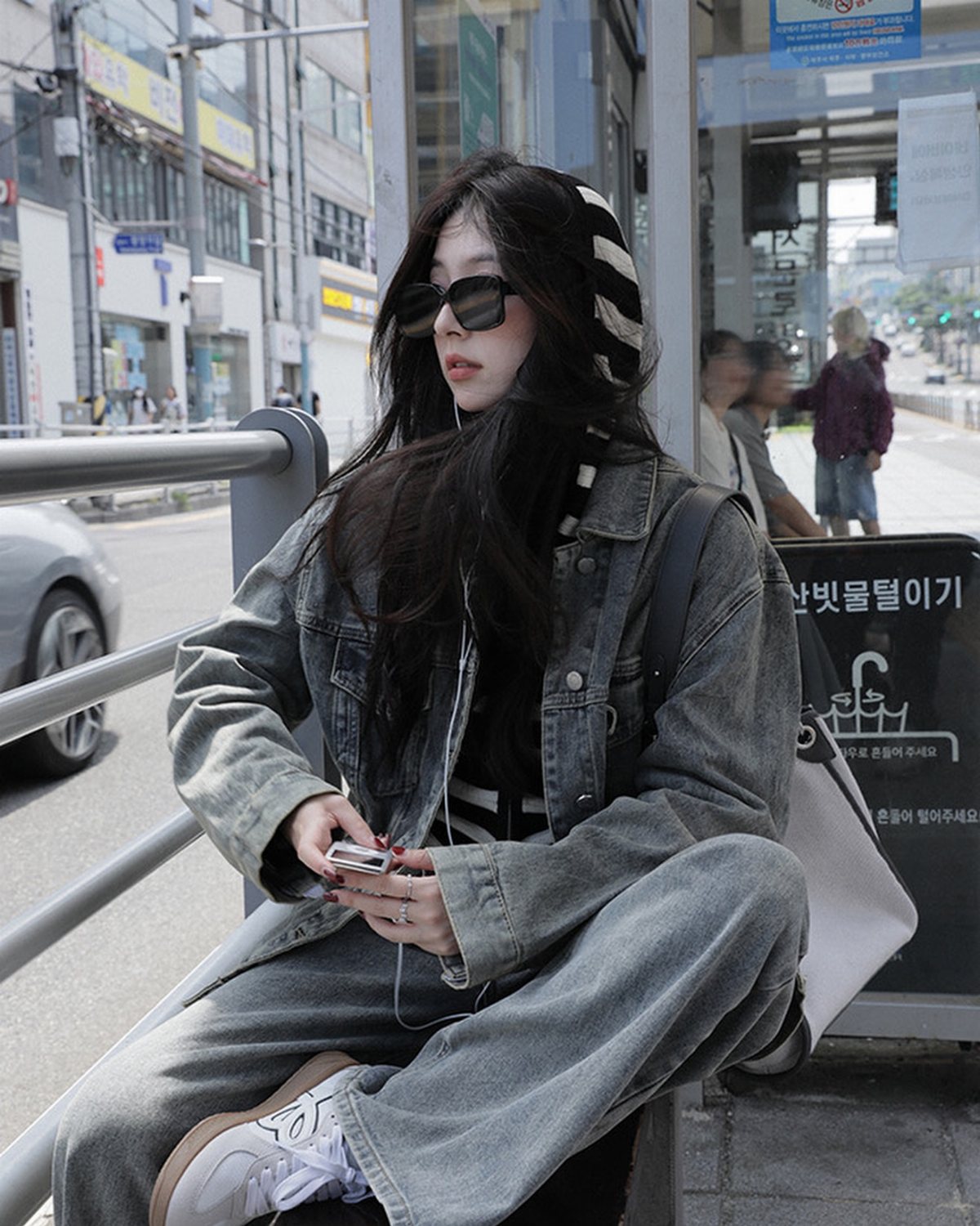 A woman wearing a denim jacket and striped hoodie sitting on a metal railing outdoors.