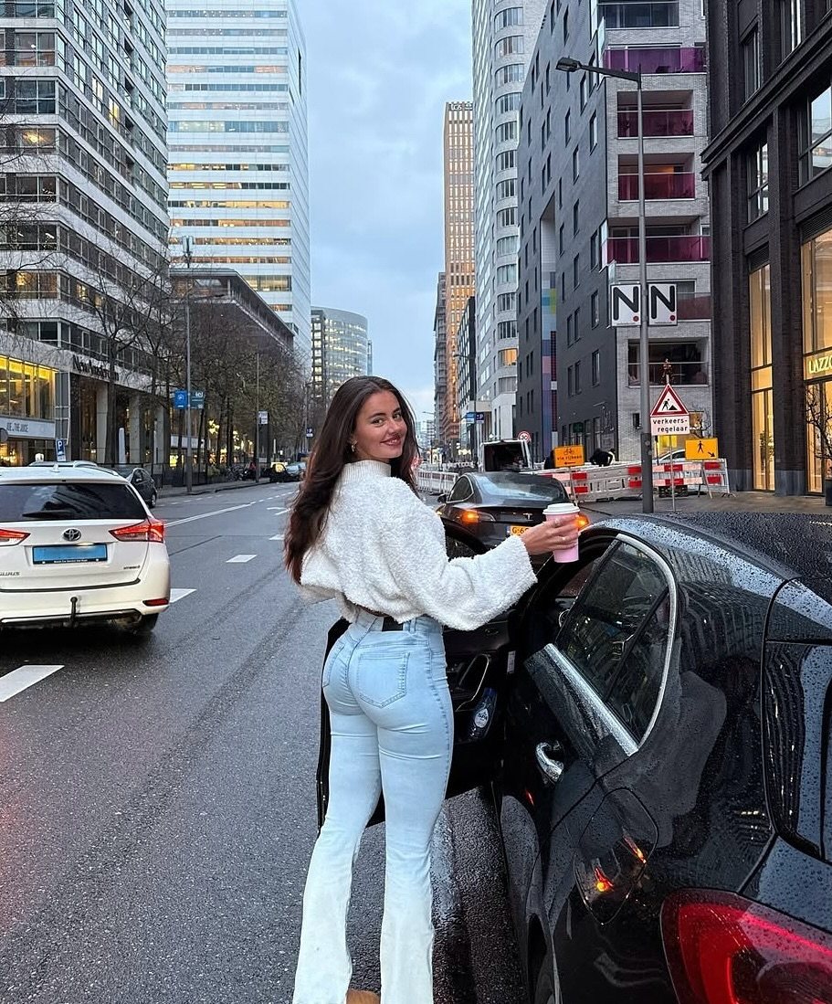 A woman in a white sweater and light blue flare jeans standing next to a black car.