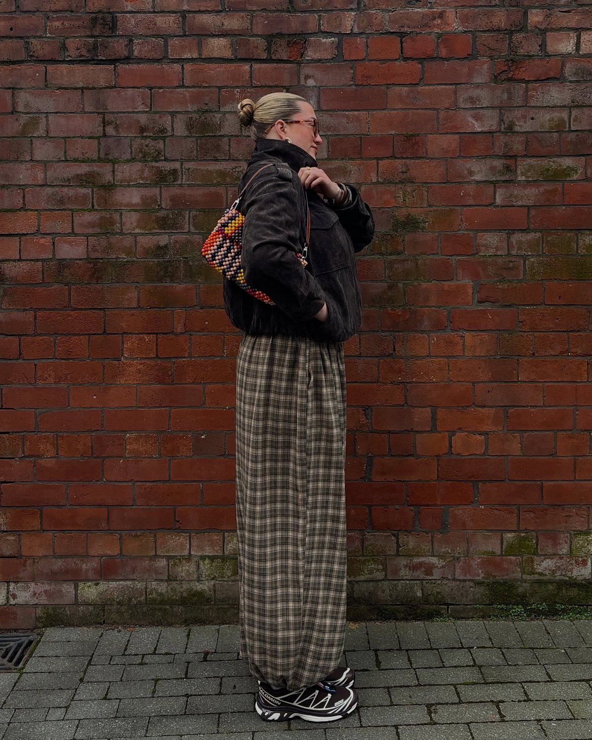 A woman in a dark jacket and long plaid skirt stands against a brick wall.