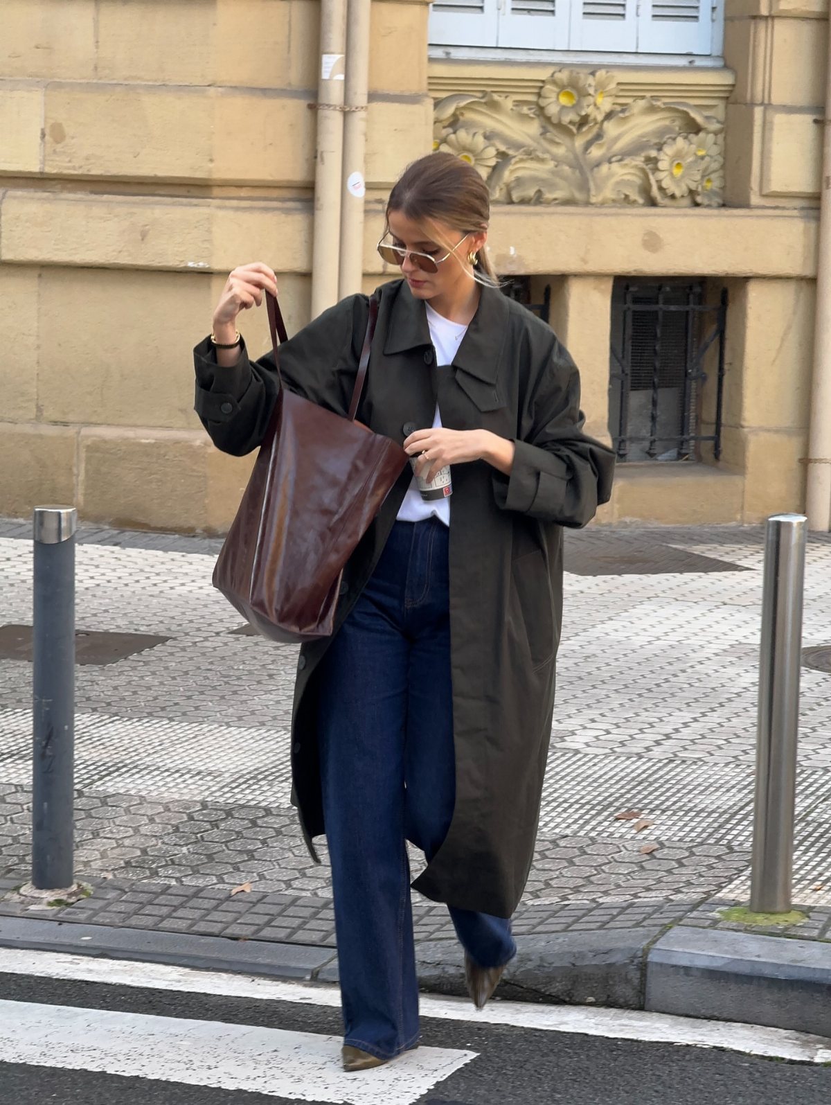 Woman in a long olive trench coat and blue jeans walking through a city street.