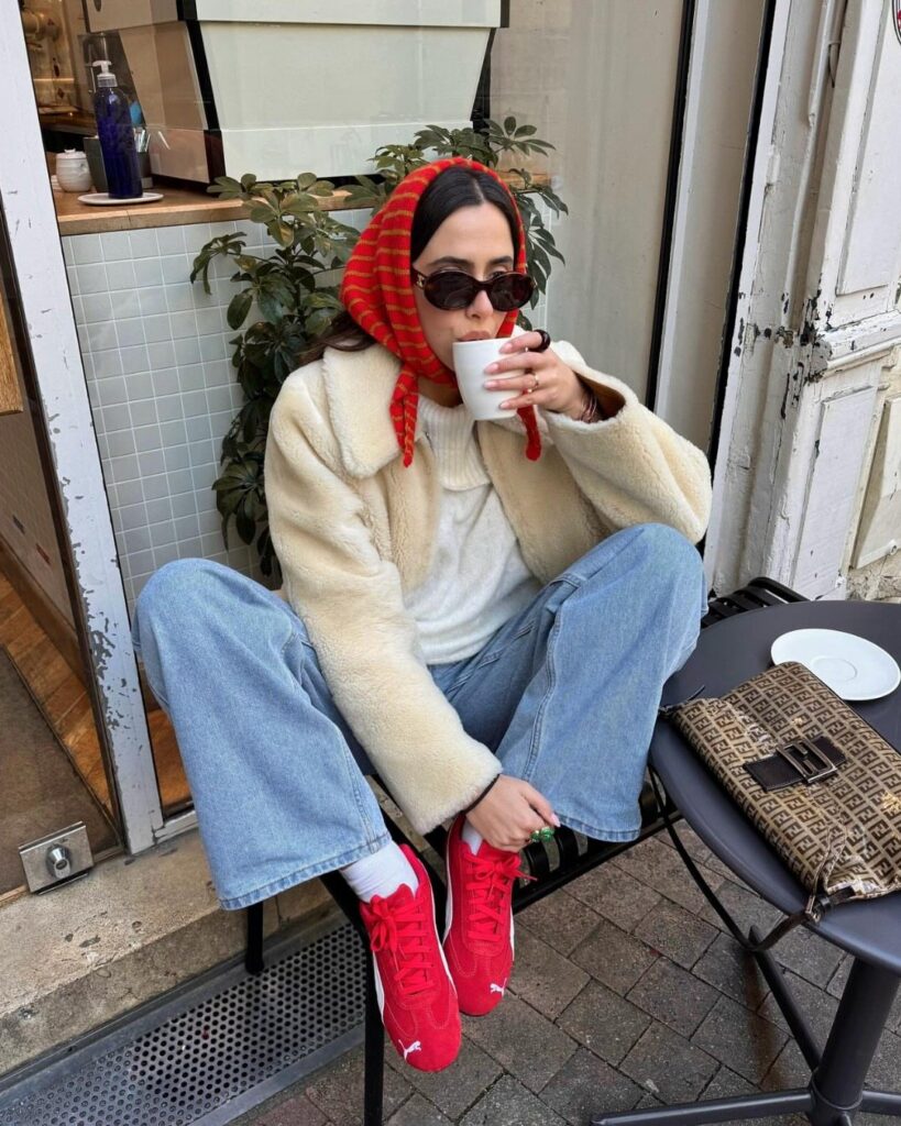 Woman in cream jacket, blue jeans, and red sneakers sitting outdoors drinking from a cup.