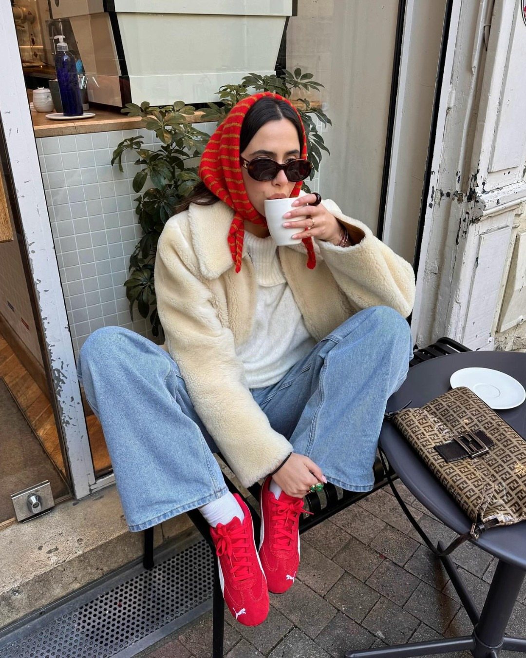 Woman in cream jacket, blue jeans, and red sneakers sitting outdoors drinking from a cup.