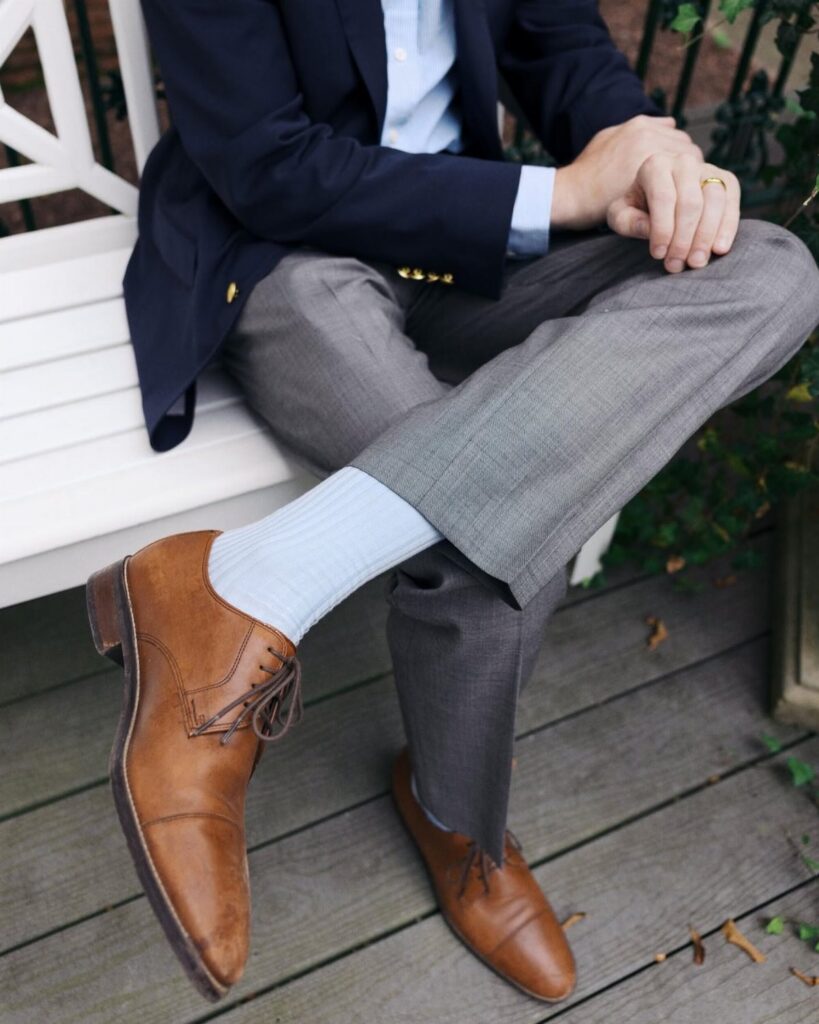 Man sitting on a white bench wearing a navy blazer, grey trousers, and light blue socks.