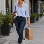 Woman walking on a sidewalk wearing a striped shirt, slim dark jeans, and white sneakers.