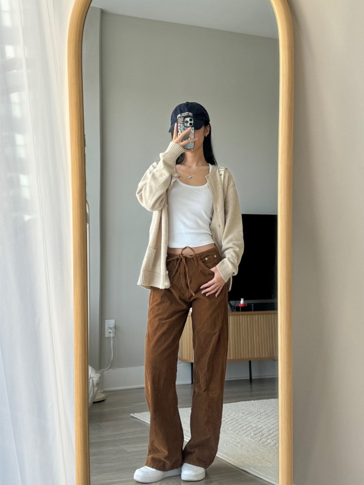 Woman in a white tank top, cream cardigan, and brown trousers posing in a mirror.