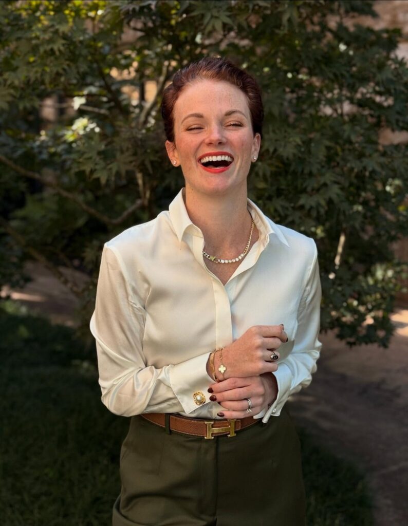 A smiling woman wearing a white silky blouse and olive green trousers outdoors.