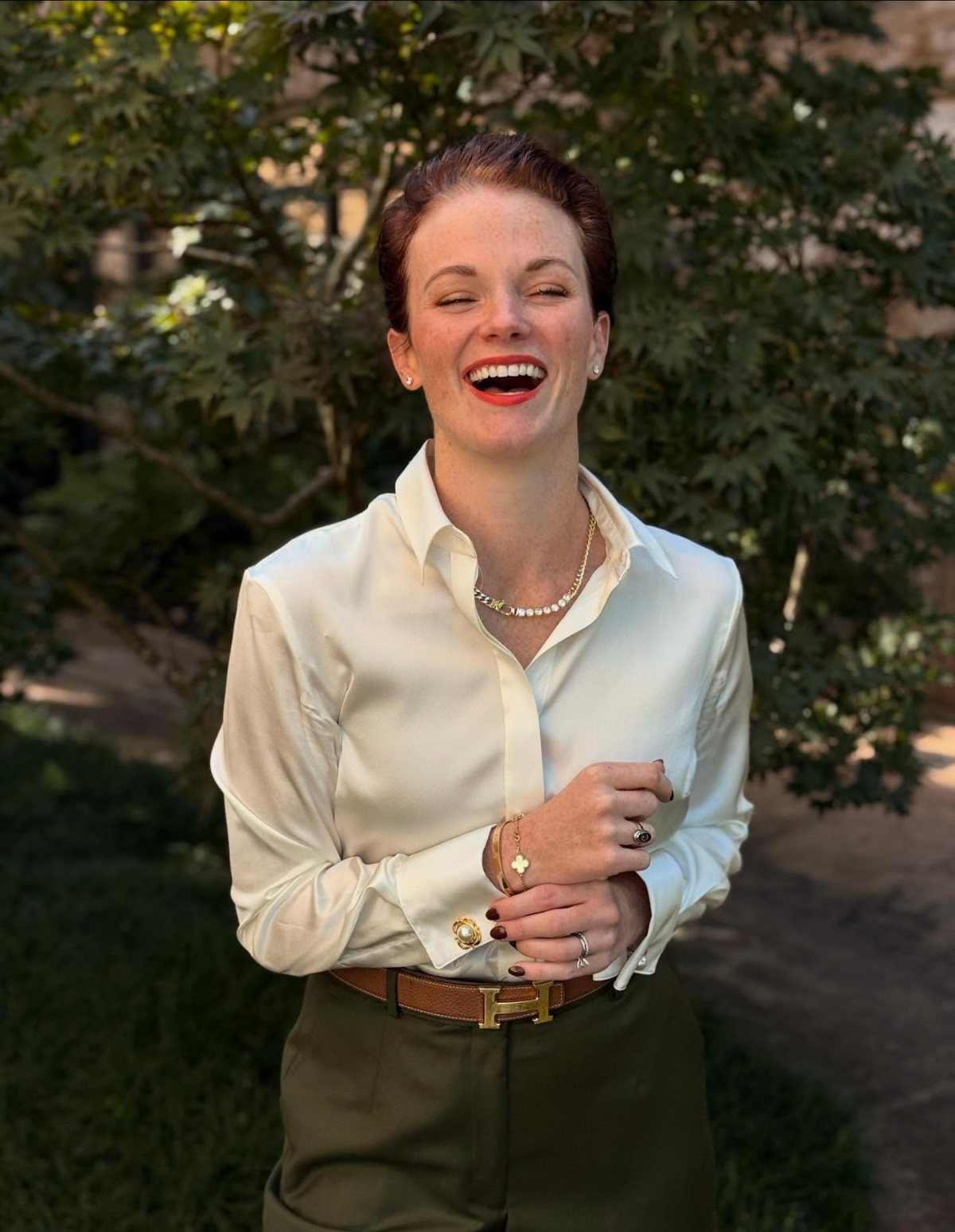 A smiling woman wearing a white silky blouse and olive green trousers outdoors.
