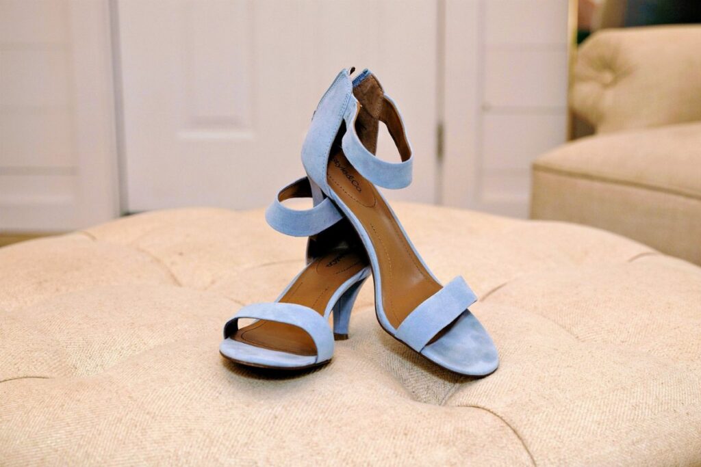 Light blue suede high-heeled sandals with ankle straps resting on a beige tufted ottoman.