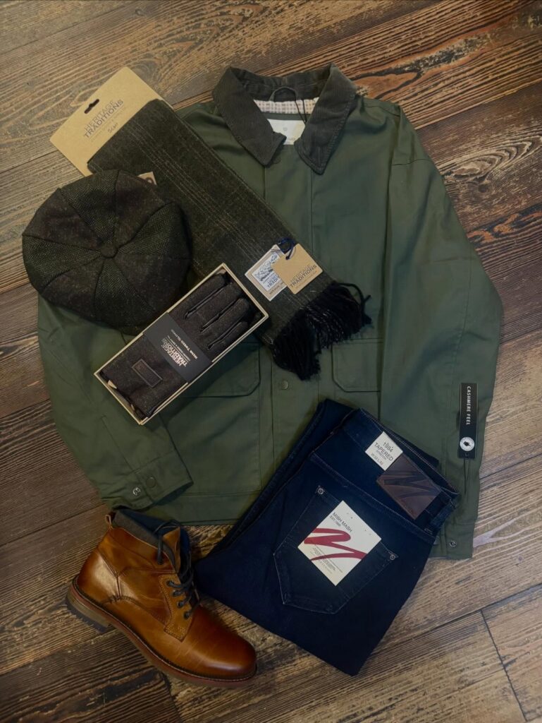 An olive green utility jacket, dark jeans, brown boots, and accessories laid on a wooden floor.