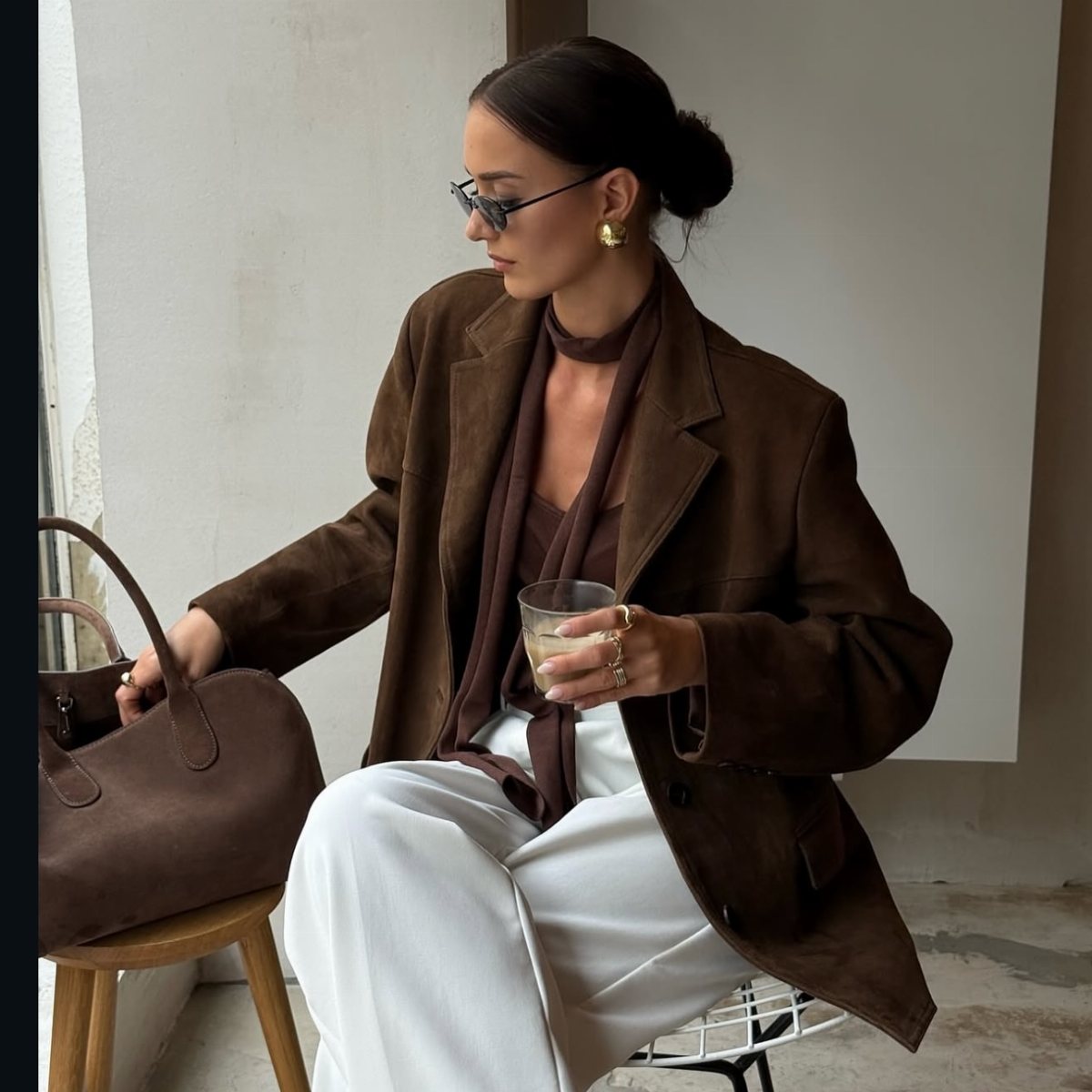 A woman in a brown suede jacket, brown scarf, and white pants sits holding a glass.