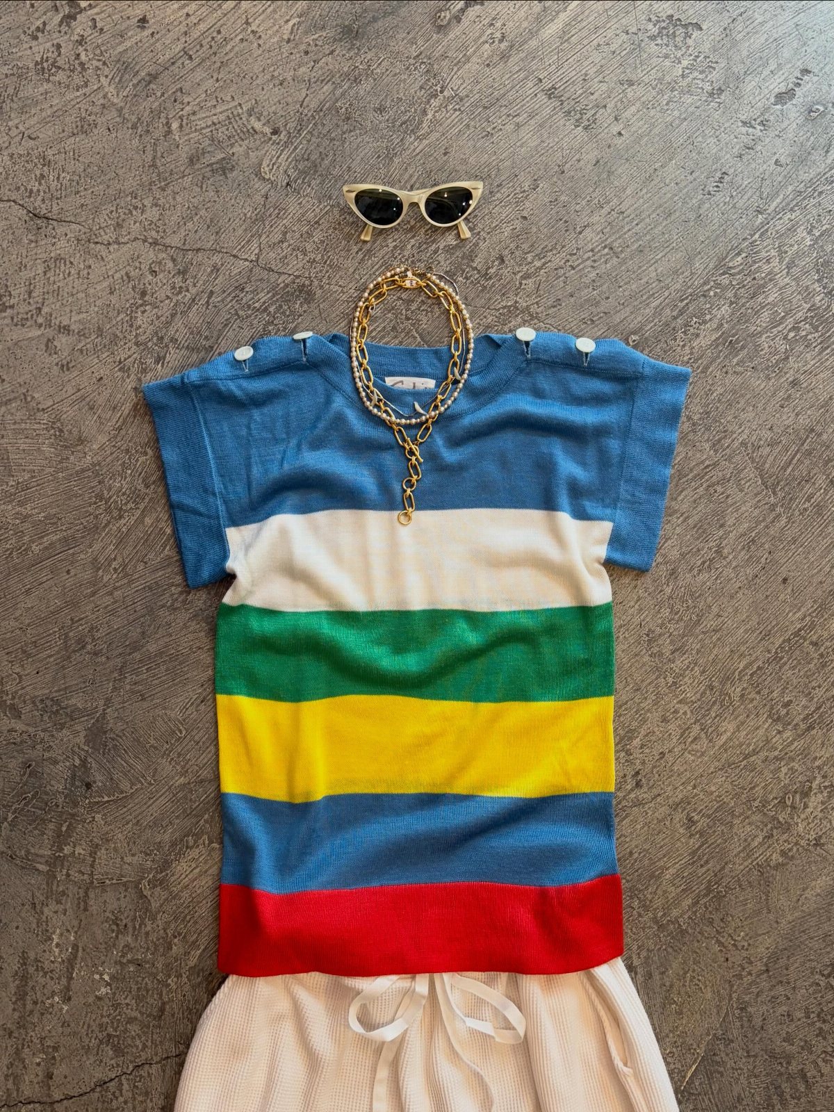 A colorful striped knit top laid flat with gold jewelry and sunglasses on a grey background.