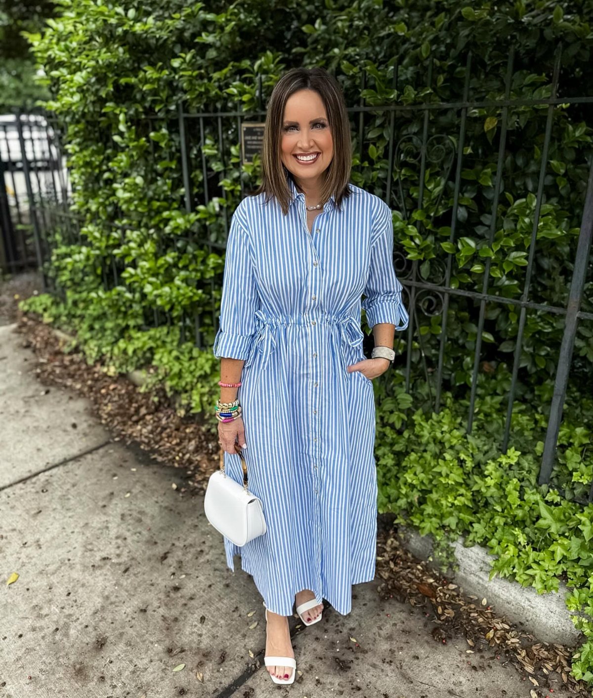 Woman in a blue and white striped midi shirtdress posing by a green hedge.