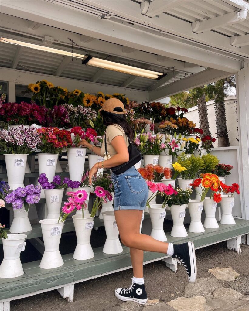A woman in denim shorts and a beige top posing next to colorful flowers.