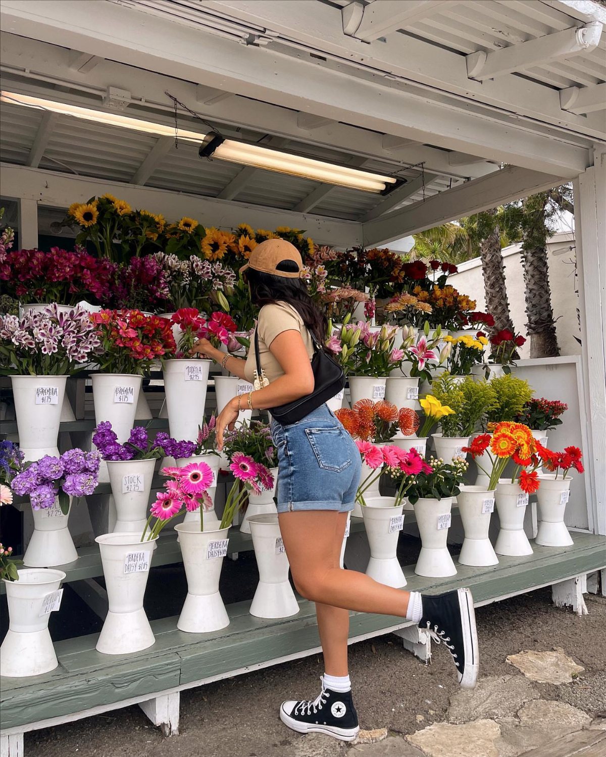 A woman in denim shorts and a beige top posing next to colorful flowers.