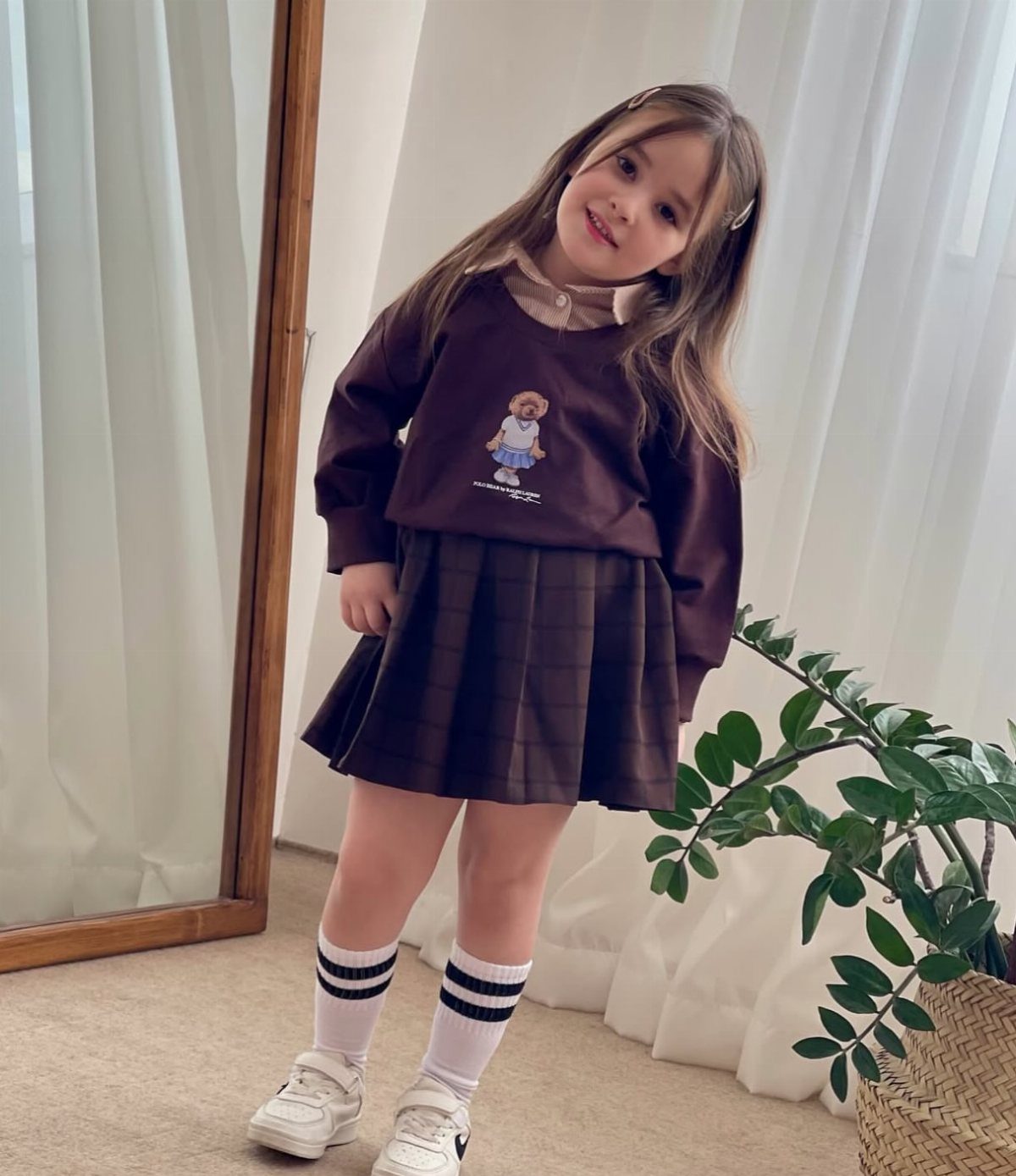 A young girl posing in a brown sweatshirt, pleated skirt, striped socks, and white sneakers.