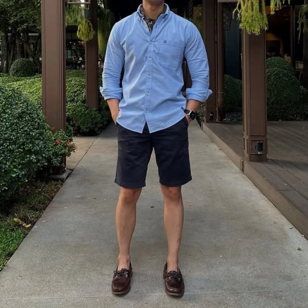 A man wearing a light blue shirt, navy shorts, and brown boat shoes standing on a walkway.