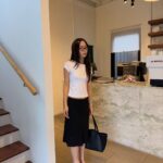 Woman standing in a room wearing a white t-shirt and a black midi skirt.