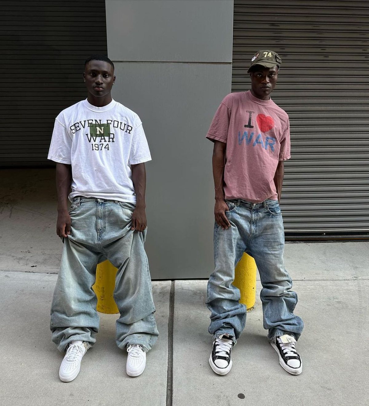 Two men standing in streetwear outfits against a grey wall and closed metal shutters.