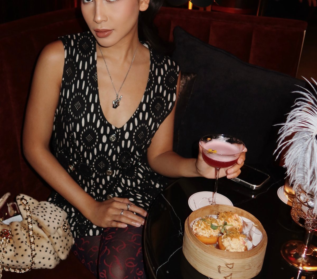 Woman in a patterned black and white top holding a pink cocktail in a lounge setting.