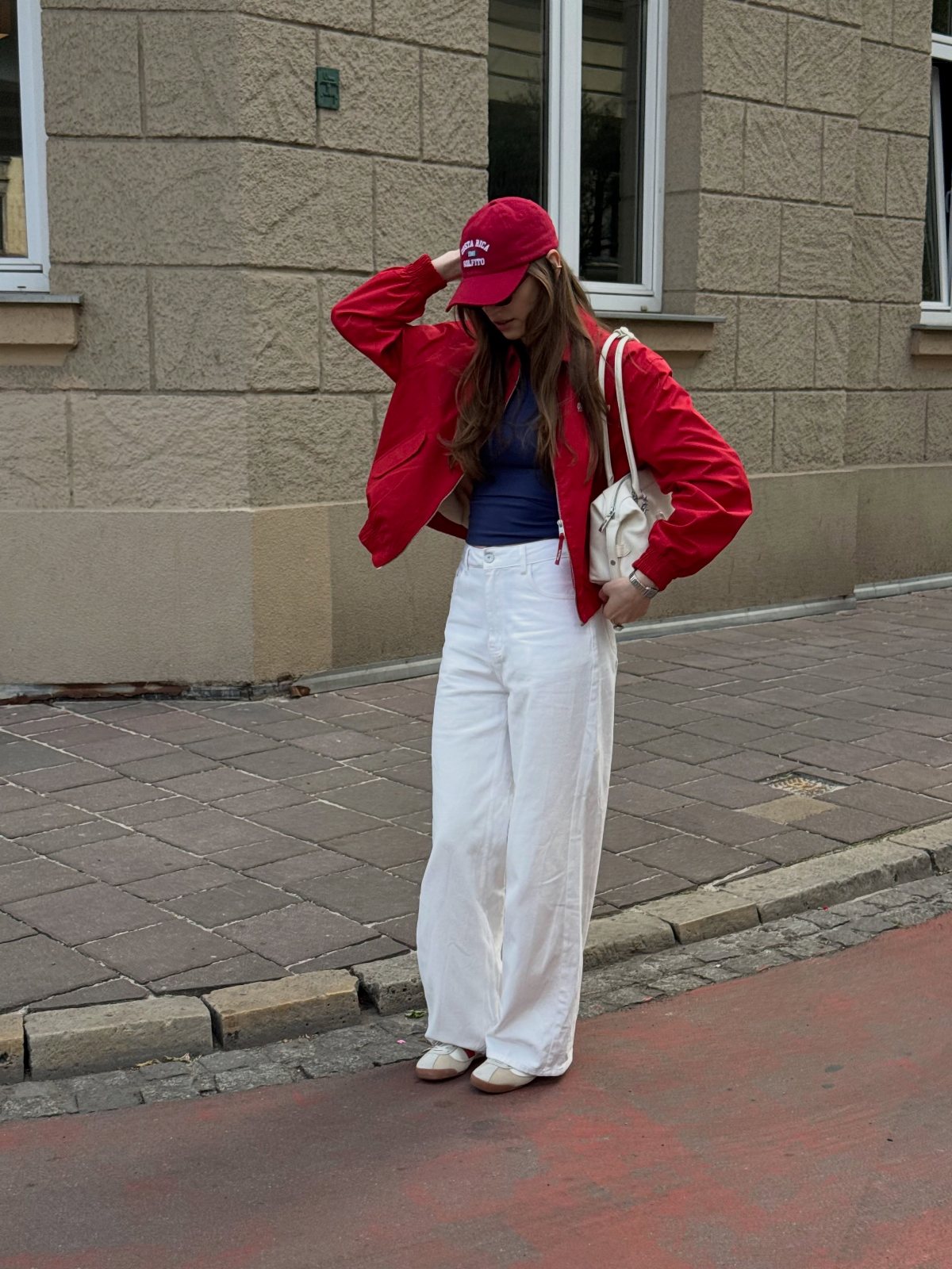 Woman in a red jacket, blue top, and white wide-leg trousers standing on a sidewalk.