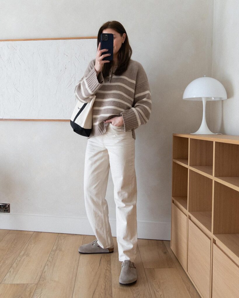 Woman wearing white jeans, a striped sweater, and grey shoes standing in a room.