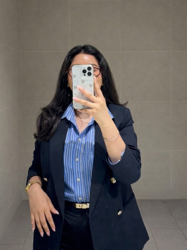 Woman in a black blazer and striped blue shirt taking a mirror selfie.