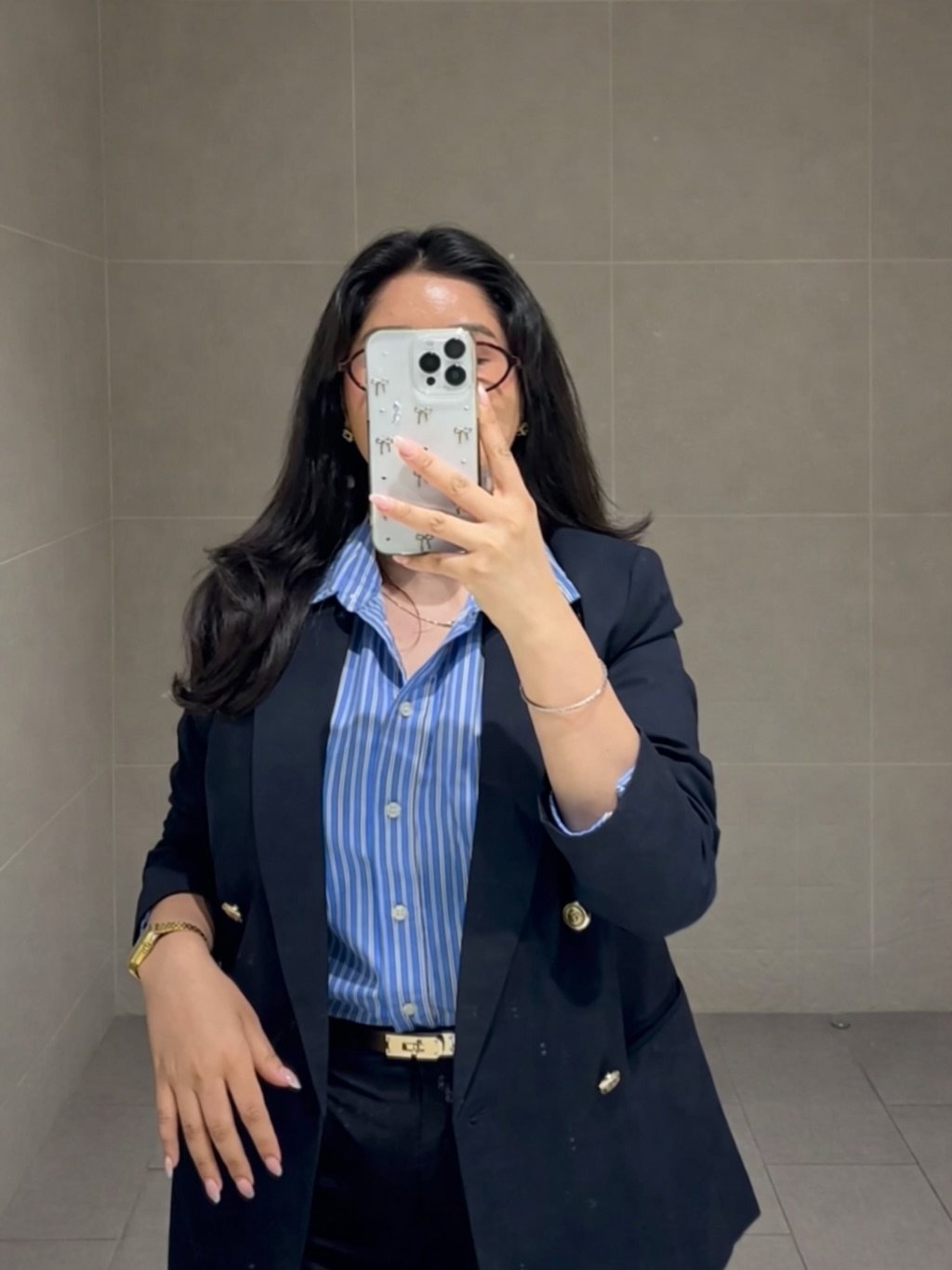 Woman in a black blazer and striped blue shirt taking a mirror selfie.